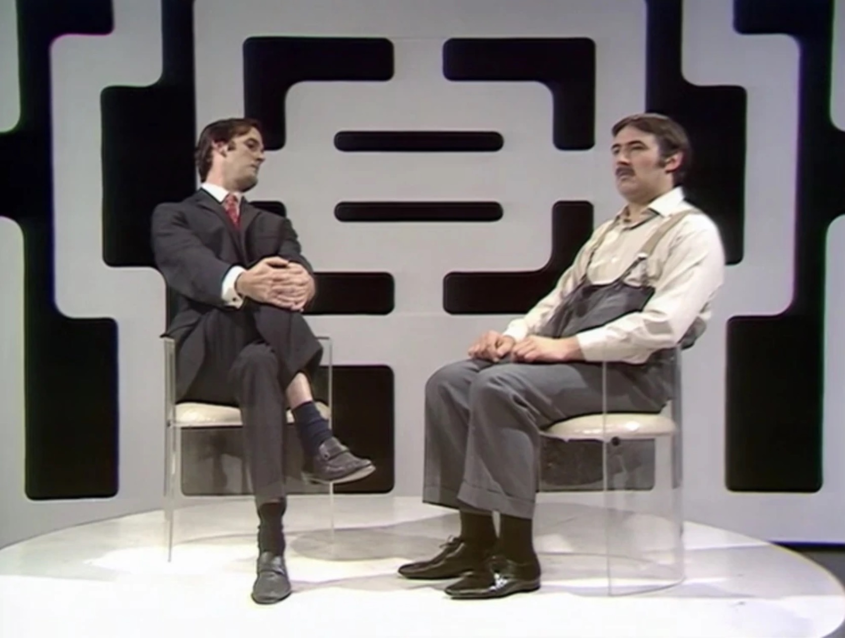 A television interviewer sits with his legs crossed away and hands clasped, eyes fixed on the exit, beside a patient man in workman's overalls who waits to be asked — John Cleese interviewing Arthur Frampton.